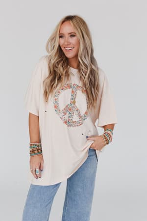 A smiling woman with long, wavy blonde hair wears the Jude Oversized Peace Sign Graphic Tee in oatmeal, paired with light blue jeans and colorful beaded bracelets, standing against a plain white background.