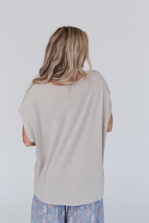 A person with long, wavy blond hair stands with their back to the camera, wearing the Camden Lace V Neck Top in camel and blue floral patterned pants against a plain light background.