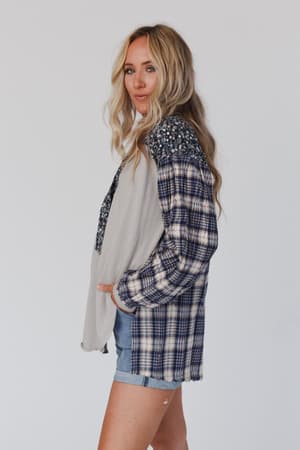 A woman with long blonde hair stands sideways against a plain white background, wearing the Better Together Mixed Print Top - Blue Gray with plaid sleeves and floral details, paired with light blue denim shorts.