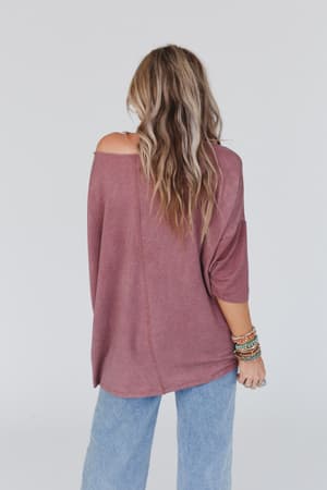 A woman with long wavy hair stands facing away, wearing The Nest Sunday Stroll Oversized Tee in mauve, styled off one shoulder with light blue jeans and colorful bracelets, against a plain light background.