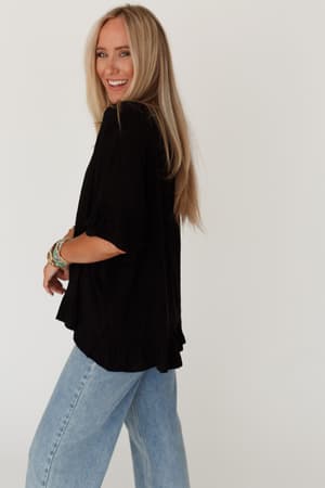 A smiling woman with long blonde hair wears The Nest Let Me Be Ruffled Top in black and light blue jeans, standing sideways and looking back at the camera against a plain white background. She accessorizes with bracelets.