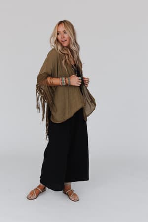 A woman with long, wavy blonde hair wears the Dreamy Macramé Kimono in olive over a black jumpsuit, paired with sandals and colorful beaded bracelets, standing against a plain white background.