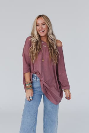 A woman with long blonde hair is smiling and standing against a plain light background, wearing The Nest Sunday Stroll Oversized Tee in mauve, paired with light blue jeans and several colorful bracelets.