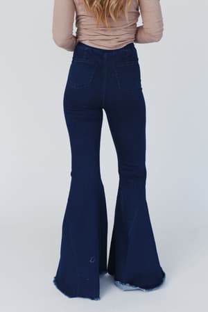 A person with long, wavy hair is seen from the back wearing The Signature Flare - Dark Denim jeans and a beige long-sleeve top, set against a plain white background.