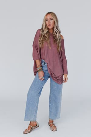 A woman with long wavy blonde hair stands against a plain background in a relaxed pose, wearing The Nest Sunday Stroll Oversized Tee in mauve, light blue wide-leg jeans, tan sandals, and colorful bracelets.