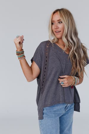 A woman with long blonde hair wears The Nest Camden Lace V Neck Top in Charcoal, blue jeans, and colorful beaded bracelets and rings. She stands against a plain light background, looking to the side.