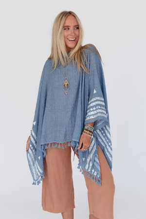 A woman with long blonde hair smiles while wearing the Fallon Embroidered Sleeve Tassel Poncho in blue, featuring white patterns and tassels. She pairs it with light brown pants and layered jewelry, standing against a plain background.