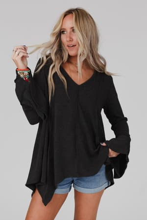 A woman with long blonde hair wears the Golden Hour Long Bell Sleeve Top in charcoal and denim shorts, standing against a plain background while holding a strand of her hair and gazing to the side.
