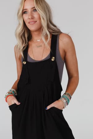 A woman with long blonde hair wears The Nest Serenity Wide Leg Jumpsuit - Black over a gray tank top, accessorized with colorful beaded bracelets and layered necklaces, posing with hands in her pockets against a plain light background.
