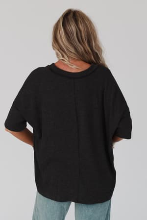 A person with long, wavy blonde hair is shown from the back, wearing The Nest On The Go V Neck Ribbed Essential Tee in Charcoal and light blue jeans against a plain light background.