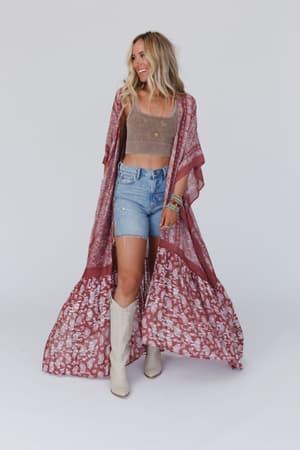 A woman wearing a paisley tapestry duster kimono in wine color with an open silhouette and kimono sleeves, paired with denim shorts.