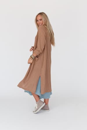 A smiling woman with long blonde hair wears The Nest Jayde Waffle Knit Cardigan in Taupe over a blue dress and gray high-top sneakers. She stands in profile, looking over her shoulder against a plain white background.
