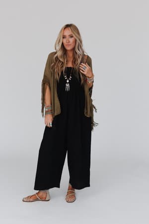 Woman with long blonde hair wears the Dreamy Macramé Kimono - Olive over a black jumpsuit, styled with sandals, layered necklaces, and multiple bracelets, standing against a plain white background.