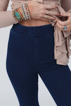 A person models The Signature Flare - Dark Denim, high-waisted stretch denim jeggings, paired with a beige long-sleeve top partially lifted. They accessorize with multiple bracelets and rings. The backdrop is plain white.