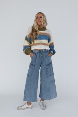 A person wearing wide leg cargo pants in a faded navy blue color with a drawstring waist and raw bottom hems, paired with a colorful sweater and white sneakers.