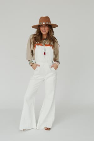 A woman stands against a plain white background, hands in her pockets, wearing a brown hat, beige long-sleeve shirt, sandals, and the Milly Distressed Hem Wide Leg Overall in Cream. She accessorizes with bracelets and layered necklaces.