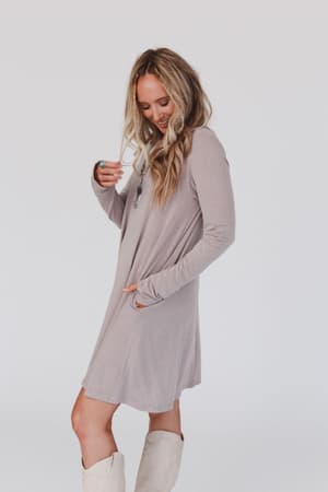 A woman with long blonde hair wears The Nest Jake Long Sleeve Crewneck Dress in Mocha Latte and white knee-high boots, standing sideways and looking down with a slight smile against a plain white background.