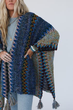 A woman with long blonde hair wears the Desert Dreams Crochet Ruana in Cobalt, featuring a textured multicolored knit with tassels over a light gray outfit. She accessorizes with rings, bracelets, layered necklaces. Plain white background; ideal for boho style.