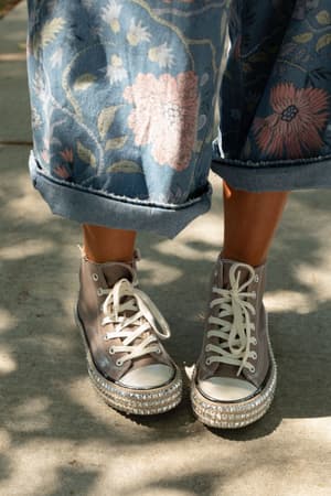 A person in floral cuffed blue jeans and Chantel Studded High Top Sneakers in Mocha, featuring thick rhinestone-embellished soles, stands on a sunlit sidewalk.