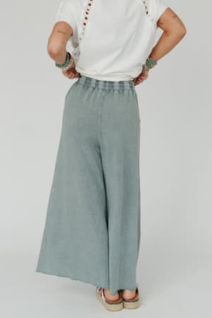 A person faces away, wearing a white eyelet short-sleeve top and the Relaxing Robin Wide Leg Pant in Faded Teal. They accessorize with bracelets and flat shoes for a comfortable, boho-inspired look.