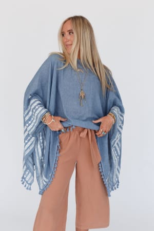 A woman with long blonde hair wears the Fallon Embroidered Sleeve Tassel Poncho in blue, featuring geometric white embroidery and tassels, over wide-legged tan pants. She accessorizes with layered necklaces and bracelets against a white background.