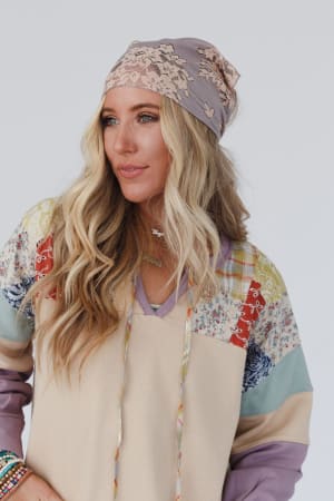 A woman with long wavy blonde hair wears the Sarah Floral Lace Headscarf in gray and a colorful patchwork hoodie. She looks to the side while standing against a plain light background.