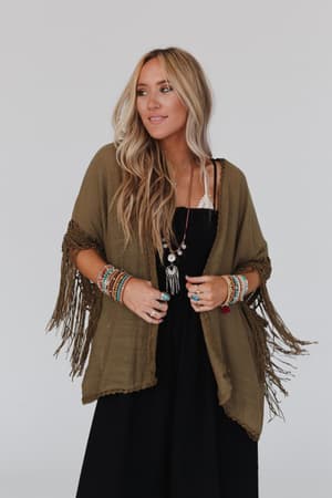 A woman with long blonde hair wears a black dress and the Dreamy Macramé Kimono in Olive, styled with colorful beaded bracelets and necklaces, standing against a plain light background.