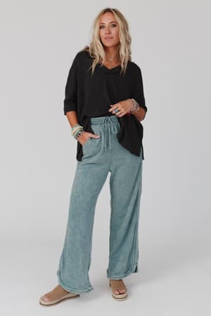 A woman with long blonde hair stands confidently against a plain background, wearing The Nest On The Go V Neck Ribbed Essential Tee in charcoal, paired with light blue wide-leg drawstring pants, tan wedge sandals, and multiple bracelets.