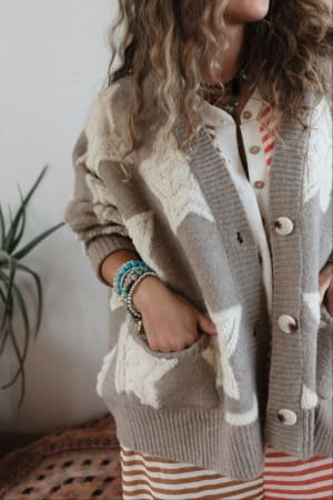 A woman with wavy hair wears the Wrapped In Stars Oversized Cardigan in taupe over a cream and red striped dress, accessorized with colorful beaded bracelets. Her hand is in her pocket, with a plant visible in the background.
