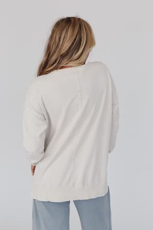 A person with long, wavy, light brown hair faces away from the camera, wearing the Catch My Smile Pullover Top in khaki and light blue pants against a plain, light-colored background.