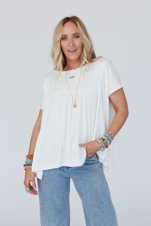 A woman with long blonde hair wears the Sunday Morning Babydoll Top in white, paired with blue jeans and colorful jewelry, standing against a plain light background and smiling softly with one hand in her pocket.
