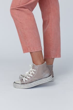 A person wears light pink jeans and Chantel Studded High Top Sneakers in Mocha, featuring thick rhinestone-detailed platforms, standing on a plain light background. Only their lower legs and feet are visible.