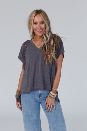 A woman with long blonde hair smiles at the camera, wearing The Nest Camden Lace V Neck Top in Charcoal and light blue jeans. She accessorizes with colorful bracelets and multiple rings. The background is plain and light-colored.