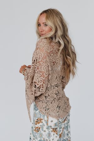 A woman with long, wavy blonde hair models the Crochet Petal Kimono Wrap in Mocha, styled over a floral skirt, standing sideways and looking over her shoulder against a plain white background.