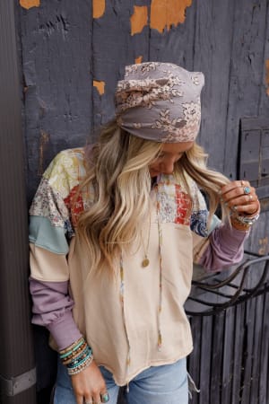 A woman with long blonde hair touches her hair while wearing the Sarah Floral Lace Headscarf in Gray, a patchwork hoodie, jeans, beaded bracelets, rings, and a pendant necklace. She stands against a textured dark wooden wall.