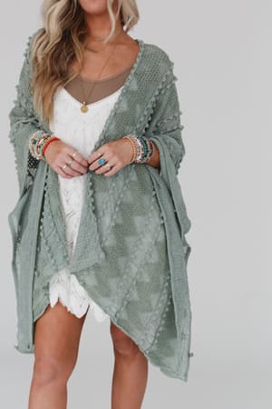 A woman models the Bali Embroidered Kimono in sage over a white dress and brown top, styled with colorful beaded jewelry. She stands against a light background, her face partially out of frame.