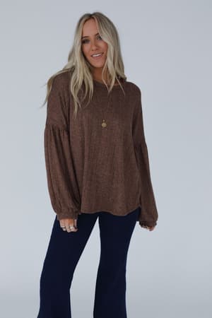A woman with long blonde hair wears The Nest Sweet Heart Balloon Sleeve Knit Sweater in Mocha with dark pants, accessorizing with rings and a gold pendant, standing against a plain light background and smiling.