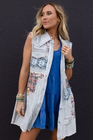 A woman with blonde hair stands against a dark wall wearing the One Of A Kind Sleeveless Denim Dress Vest - Denim Multi, featuring colorful patchwork designs, paired with multiple beaded bracelets.