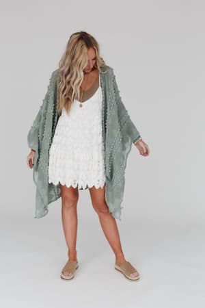 A woman with long, wavy blonde hair wears a loose white dress and the Bali Embroidered Kimono in Sage with beige sandals. She stands against a plain background, looking down with her arms slightly raised.