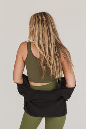 A woman with long, wavy blonde hair faces away, wearing the Hook It Up Ribbed Brami in olive and matching leggings. She drapes a black jacket over her hips against a plain, light background.