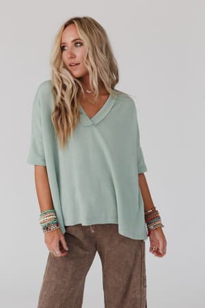 A woman with long blonde hair wears The Nest On The Go V Neck Ribbed Essential Tee in sage from Three Bird Nest, paired with brown pants and colorful beaded bracelets, standing against a plain white background.