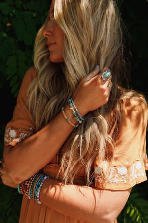 A woman with long, wavy blonde hair wears a rust embroidered dress and turquoise jewelry—including the San Antonio Bracelet Set - Turquoise—while standing outdoors against leafy greenery.