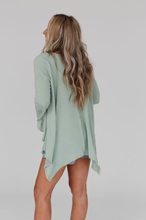 A woman with long, wavy blonde hair stands facing away, wearing the Golden Hour Long Bell Sleeve Top in sage and light denim shorts against a plain light gray background.