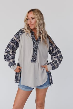 A woman with long blonde hair models the Better Together Mixed Print Top - Blue Gray, featuring floral and plaid designs, paired with denim shorts. She stands against a plain light background, looking to the side with one hand on her hip.