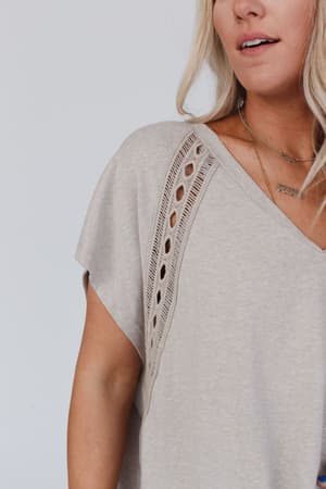 A woman wears the Camden Lace V Neck Top - Camel, featuring a knit cut-out pattern on the shoulders. With long blonde hair and layered gold necklaces, she displays her boho style. Only her upper body is visible.