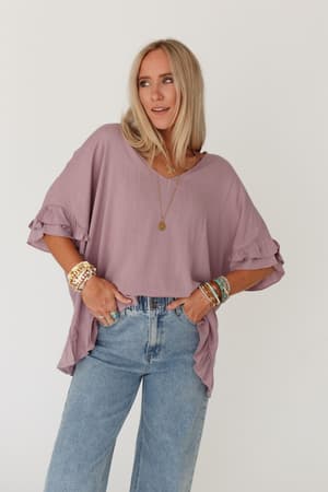 A woman with long blonde hair wears The Nest Let Me Be Ruffled Top in Pale Plum, paired with light blue jeans, layered necklaces, and colorful bracelets, standing against a plain white background with her hands in her pockets.