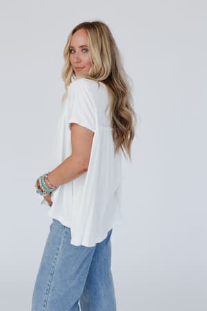 A woman with long wavy blonde hair wears the Sunday Morning Babydoll Top in white and light blue jeans, accented by colorful beaded bracelets. She stands sideways, looking over her shoulder in a relaxed boho style against a plain white background.