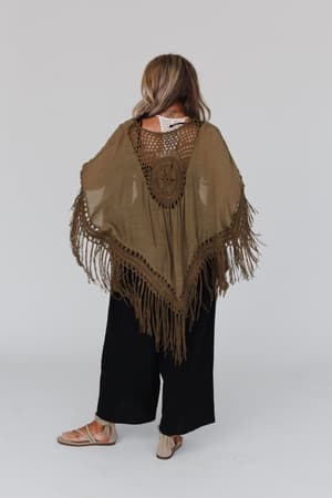 A woman stands facing away, wearing the Dreamy Macramé Kimono in olive with a circular patterned back and tassel fringe, paired with wide black pants and sandals against a plain light background.