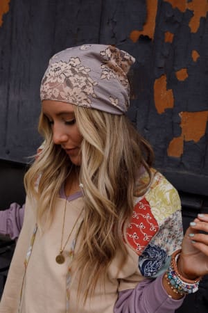A woman with long, wavy blonde hair wears the Sarah Floral Lace Headscarf in gray and a patchwork hoodie. She stands before a weathered wall, colorful bracelets on her wrist and a pendant necklace, gazing down thoughtfully.