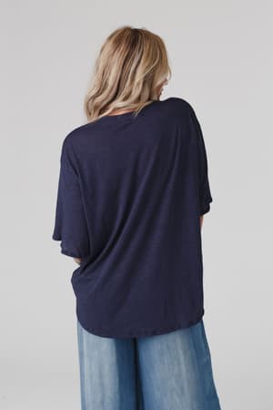 A person with long, wavy blonde hair faces away, wearing The Nest Your Favorite Slub Tee Shirt in navy and light blue wide-leg jeans against a plain light background.